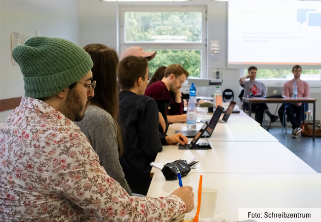 Teilnehmende an der Konferenz "Diversität im Schreibzentrumskontext" sitzen links in einer Reihe an Tischen